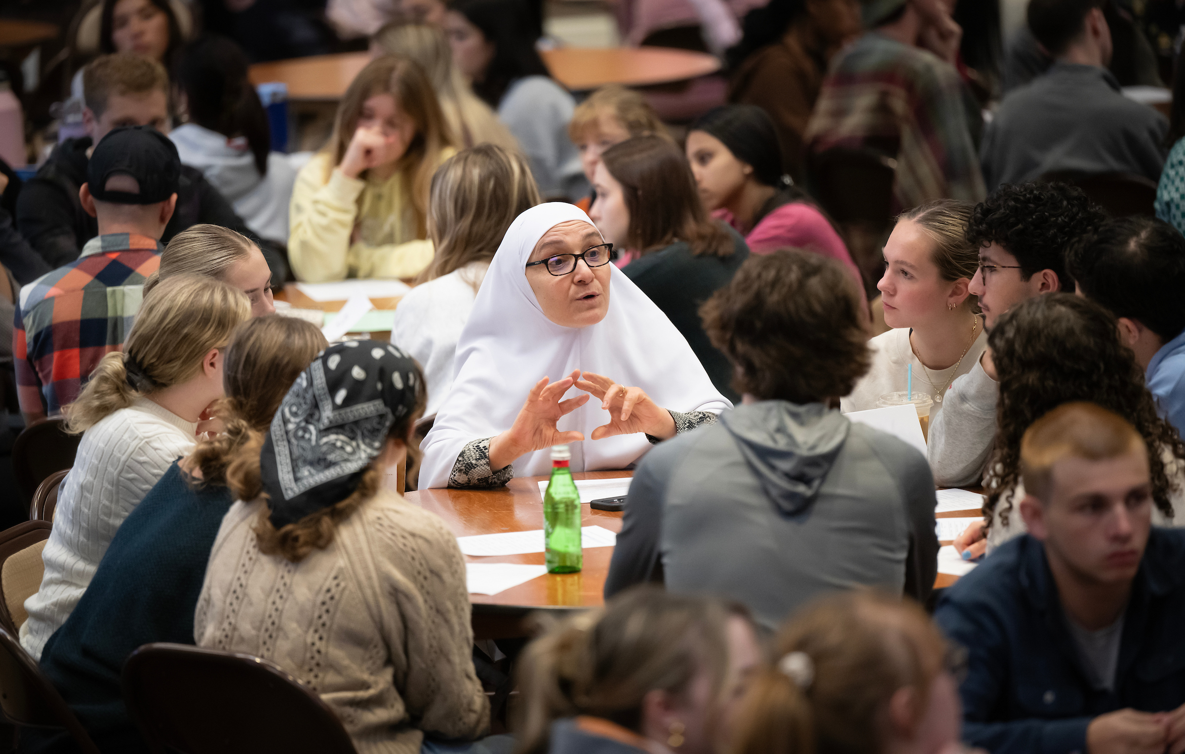 a female professor in a white hijab speaks to a circular table of students, surrounded by other tables of people conversting