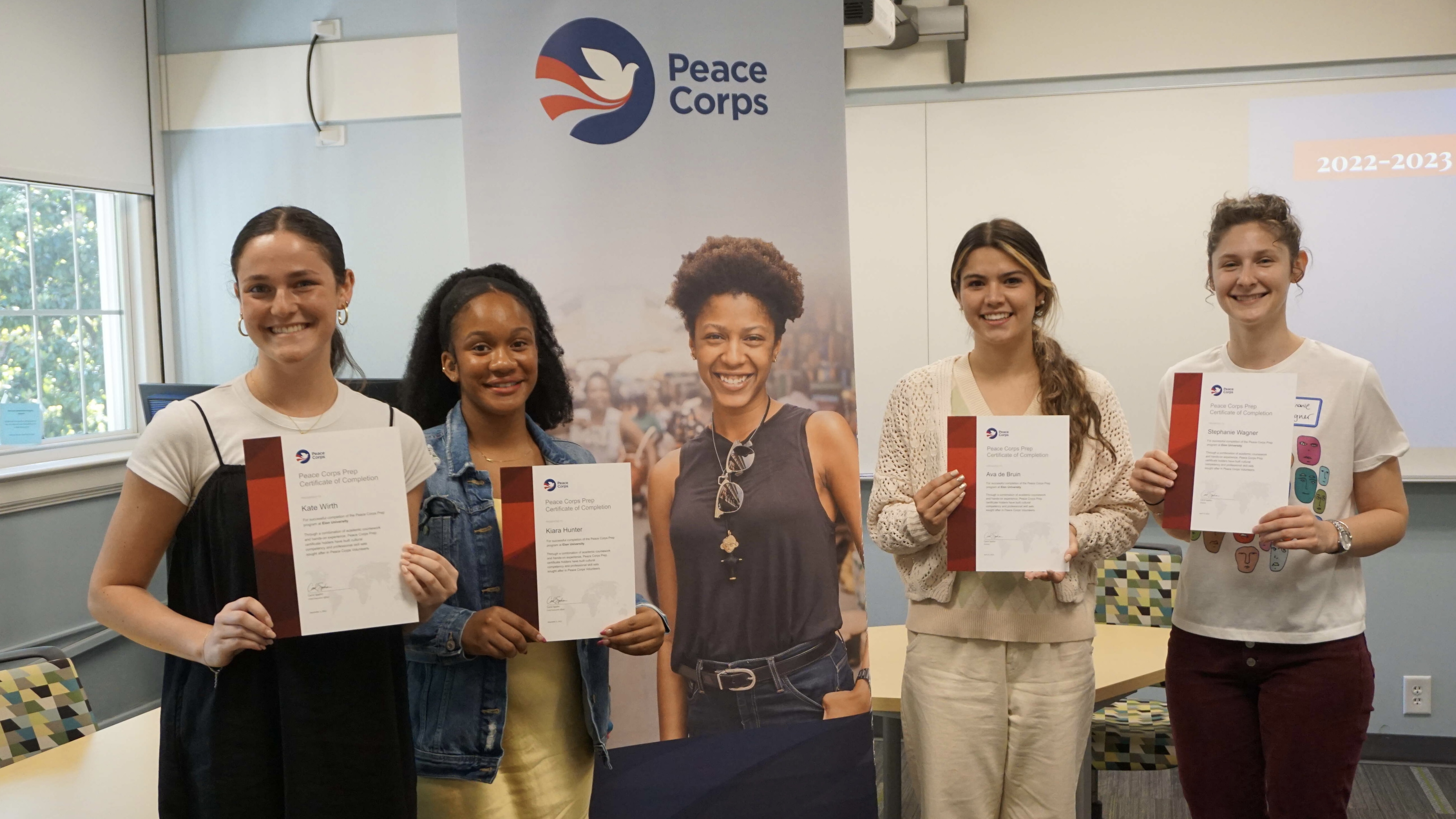 Four women students hold certificates around a vertical banner with the Peace Corps logo and photos of a volunteer 