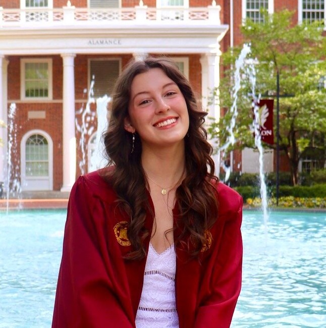 a woman in a maroon graduation gown sitting on the edge of a fountain in front of the Alamance Building