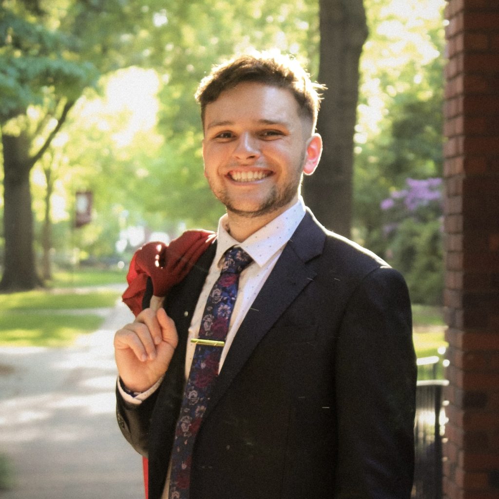 A male student in a suit with a maroon graduation gown over his shoulder standing in a brick archway