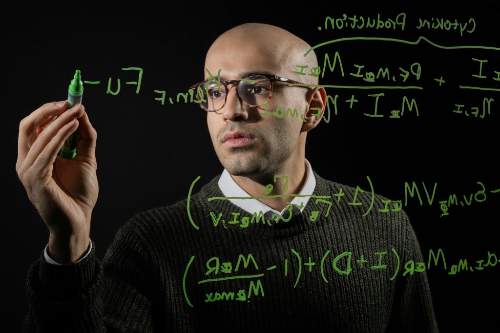 A male student writes a math problem on a transparent board using a green marker. We see his face through the board.