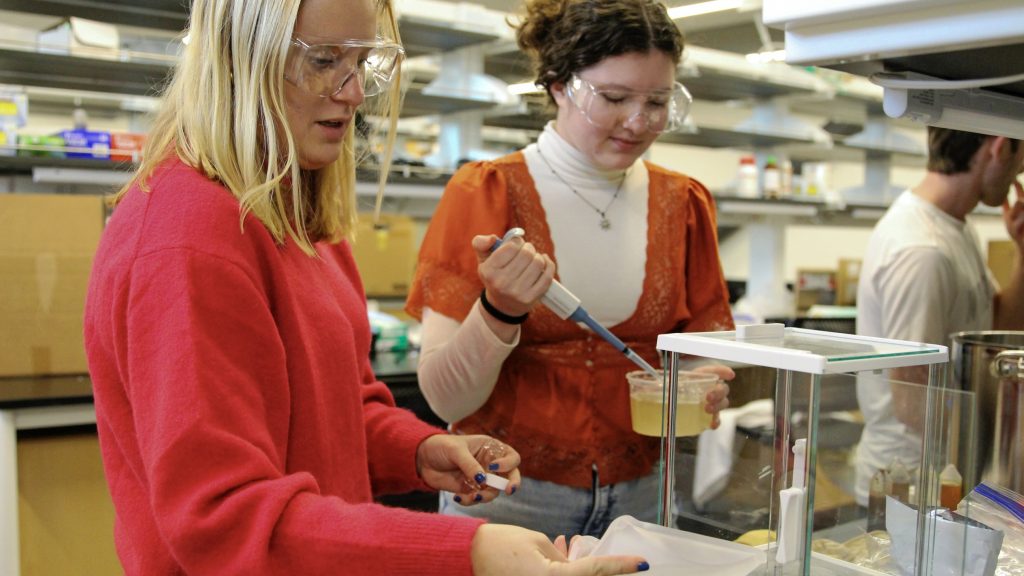 two female students wearing safety glasses prepare samples for testing in a lab using a pipette