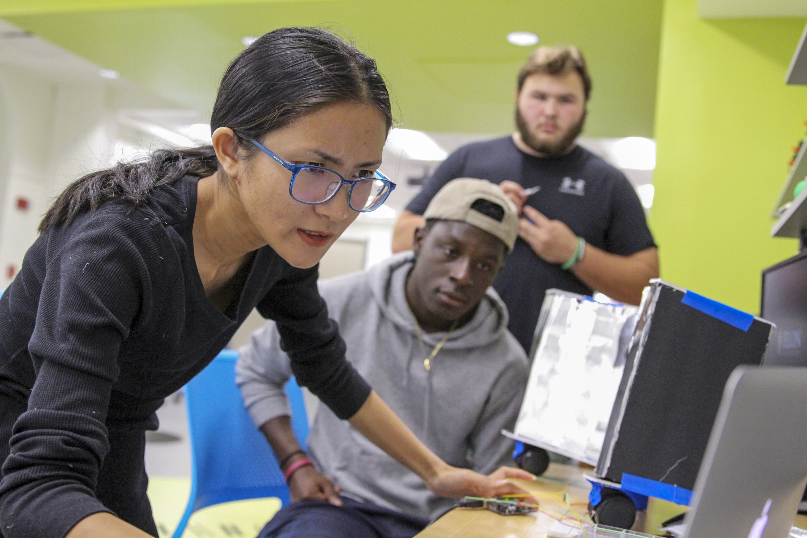 a female student wearing glasses intently works at a computer screen whiel two male students look on