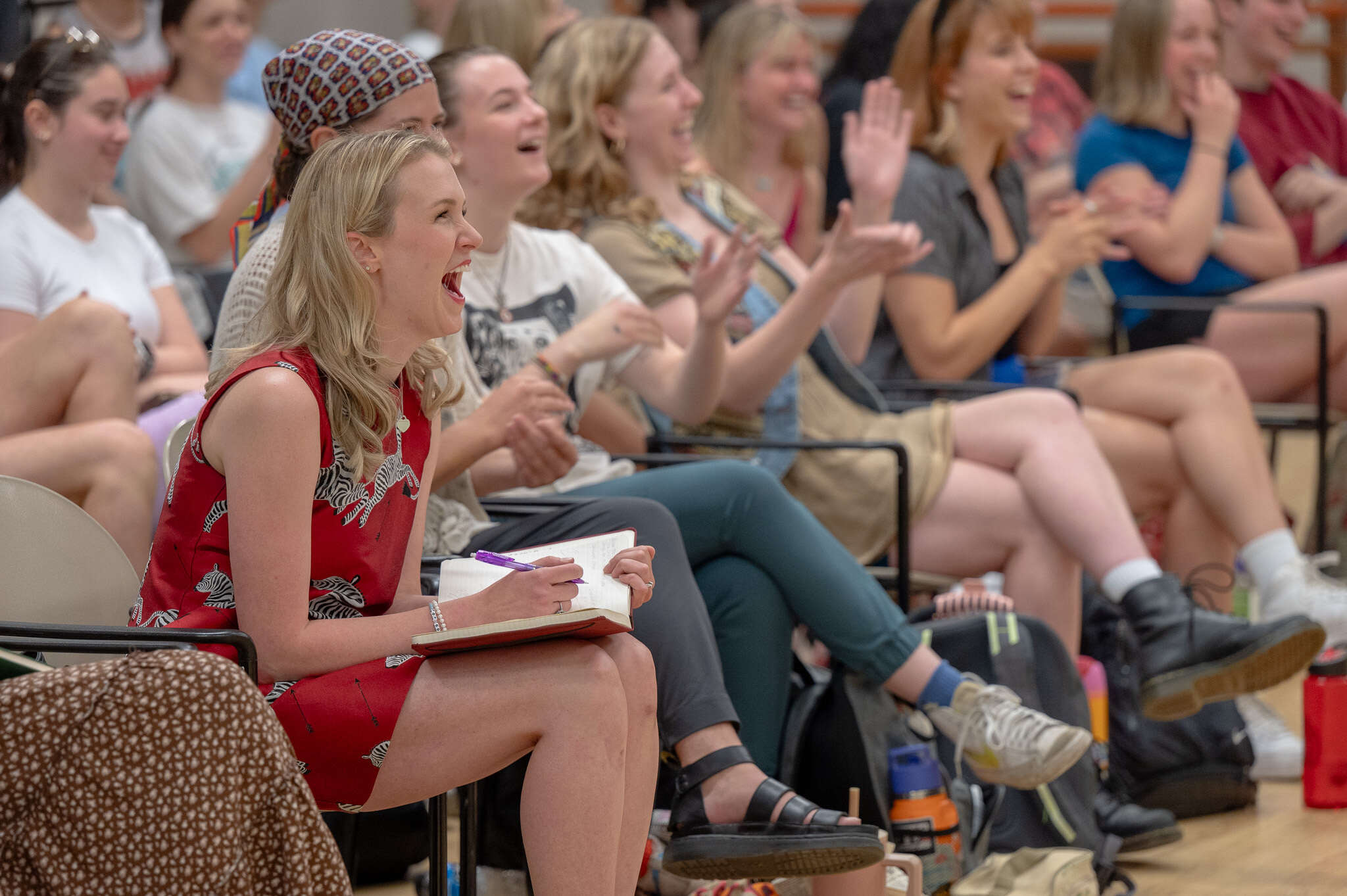 A woman with blonde hair in a red dress sits on the front of a rehearsal space with students. They are laughing and clapping. 