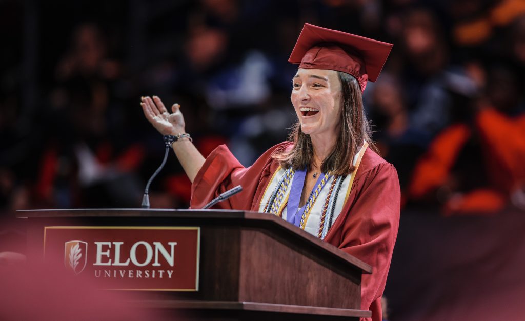 A woman in a maroon cap and gown speaks at a podium with Elon University's logo during a graduation ceremony