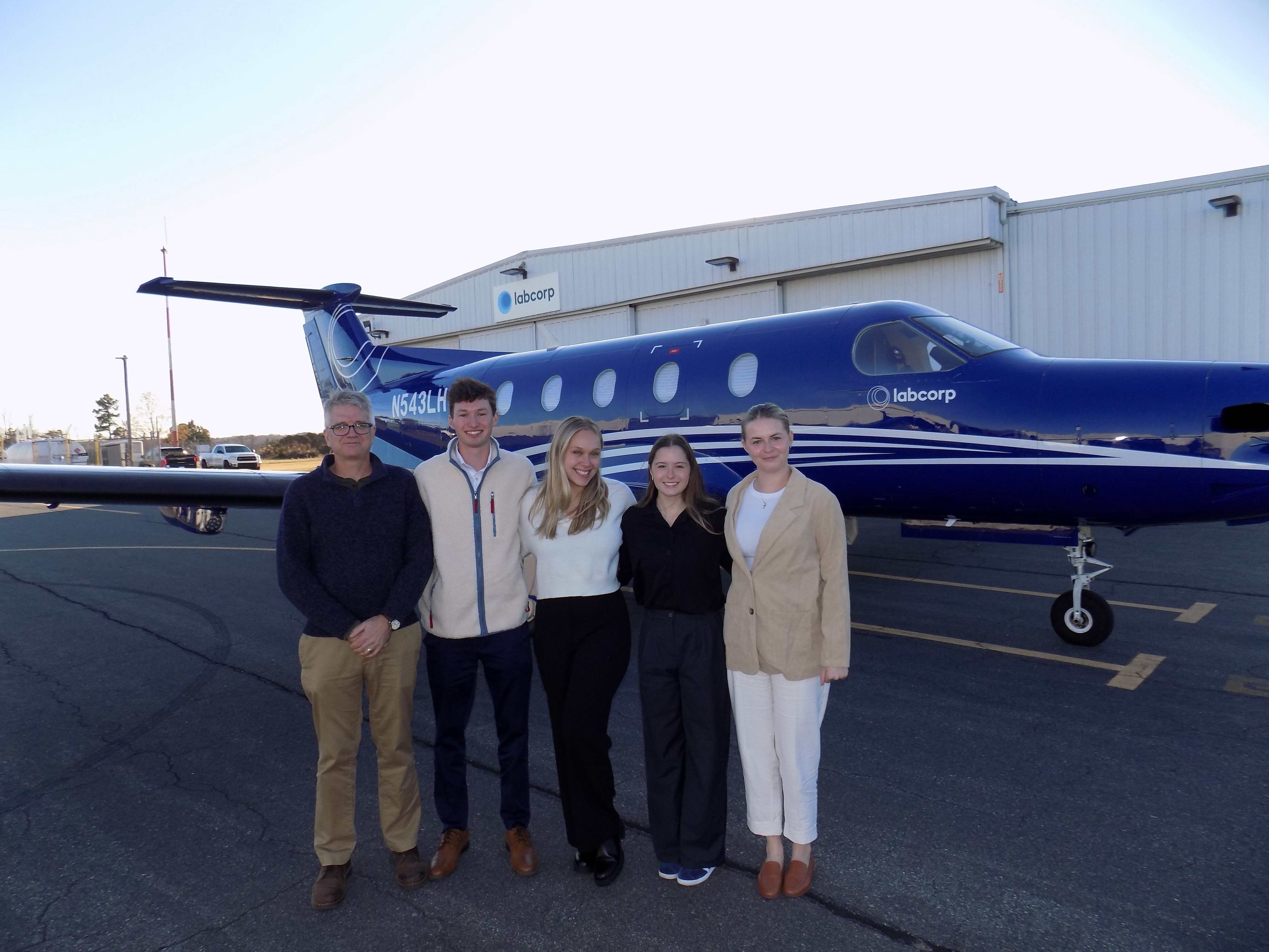 A professor and four students pose for a photo on a tarmac in front of an airplane