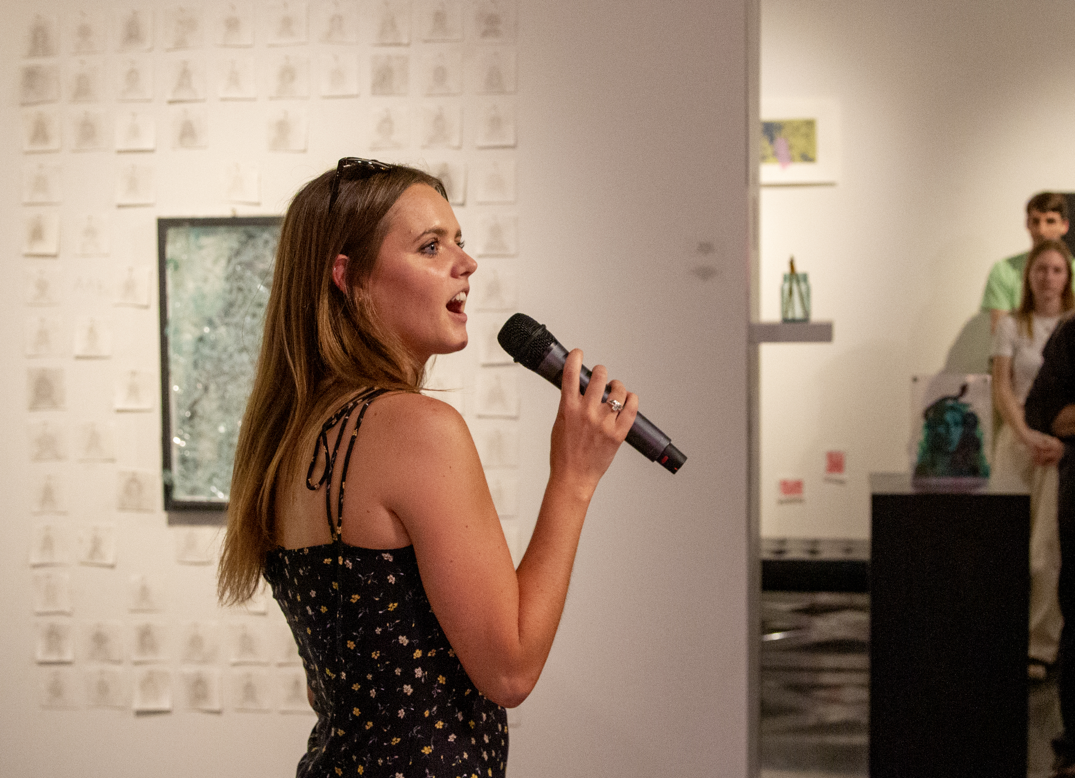a female student speaks into a microphone in an art gallery. pieces of glass art are seen behind her