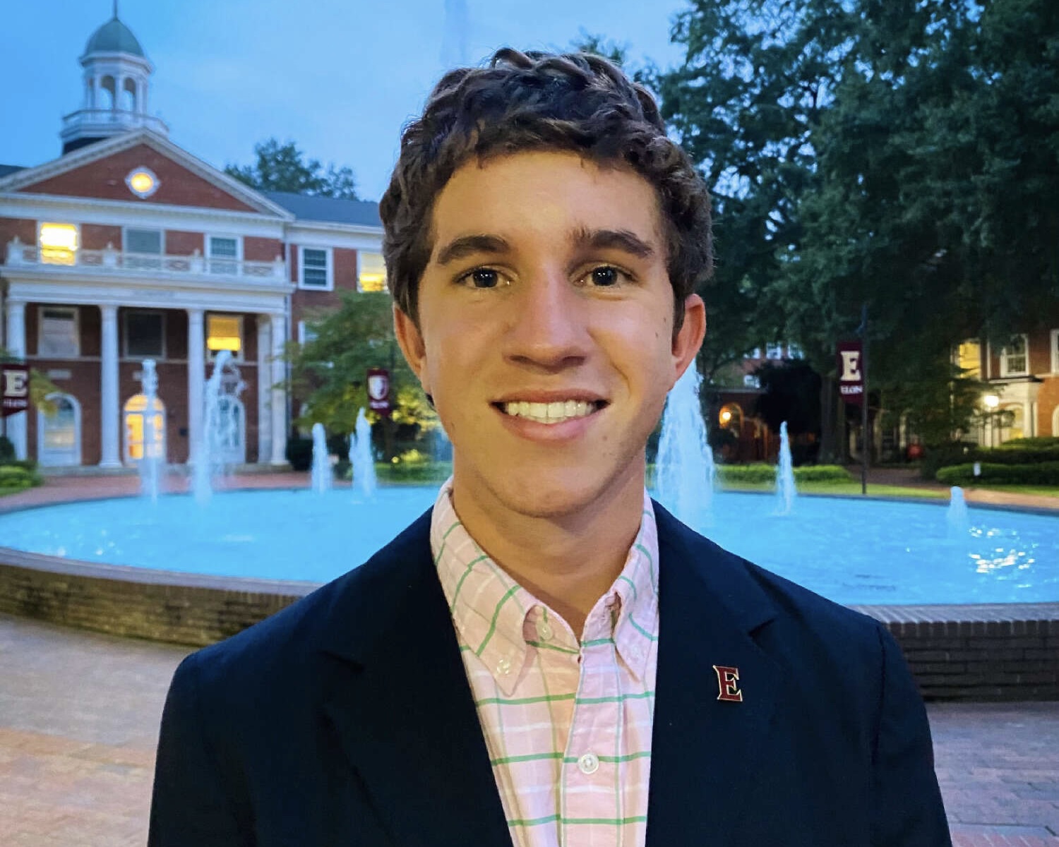 Bo Dalrymple, wearing a blue sport coat, smiles for the camera in front of Fonville fountain on Elon University's campus.