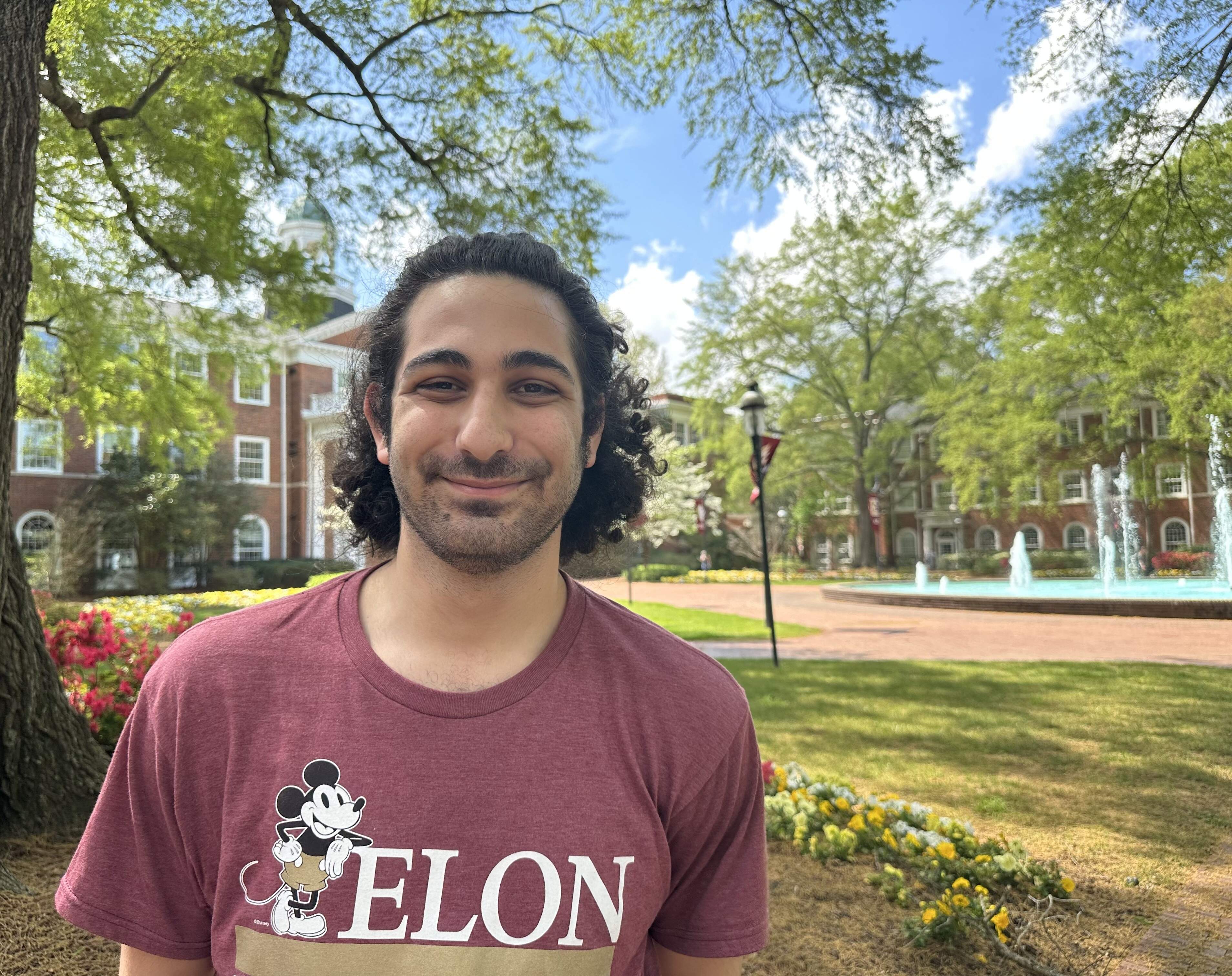 Rony Dahdal, wearing an Elon shirt, smiles for the camera in front of Fonville fountain on Elon University's campus.