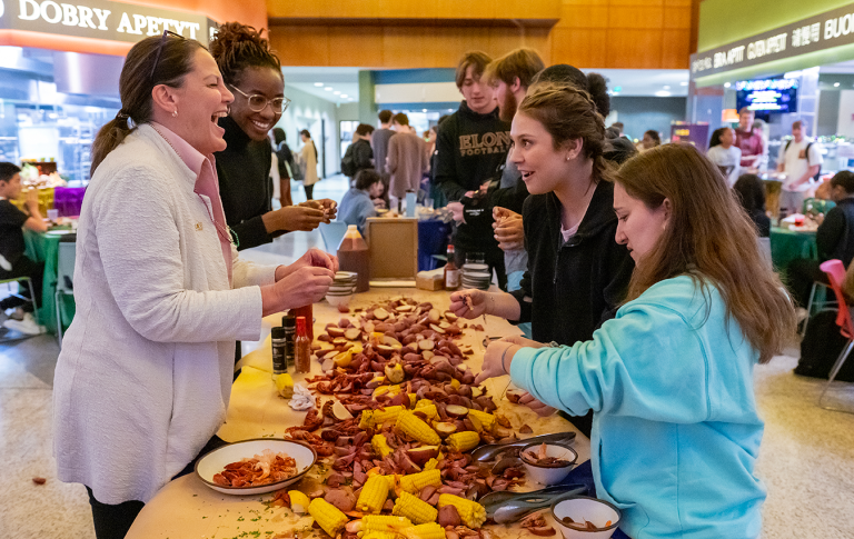 Students and staff laugh and enjoy a Mardi Gras-themed seafood boil with corn, potatoes and crawfish in a lively dining hall.