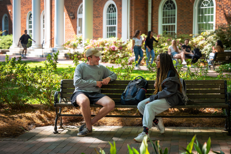 Two students sit on a bench outside a brick academic building, talking and smiling on a sunny day.