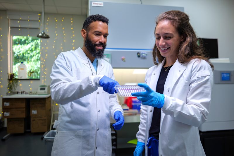 A student and a professor wearing lab coats and gloves work together on an experiment in a brightly lit science lab.