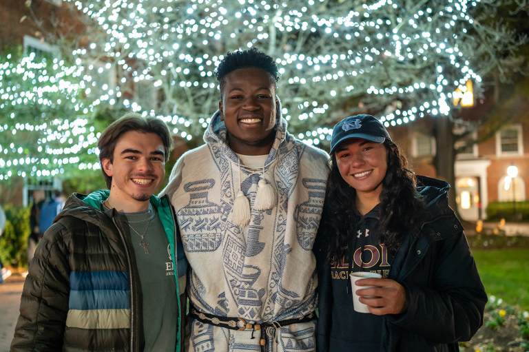 Three students pose and smile in front of twinkling holiday lights during Elon’s Festival of Lights celebration.