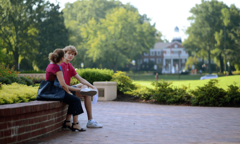 Two people sit on a brick ledge near Elon’s campus lawns, talking on a sunny day.