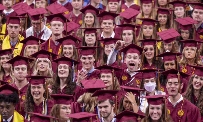 Graduating students in maroon caps and gowns gather together, smiling and cheering during Elon’s Commencement ceremony.