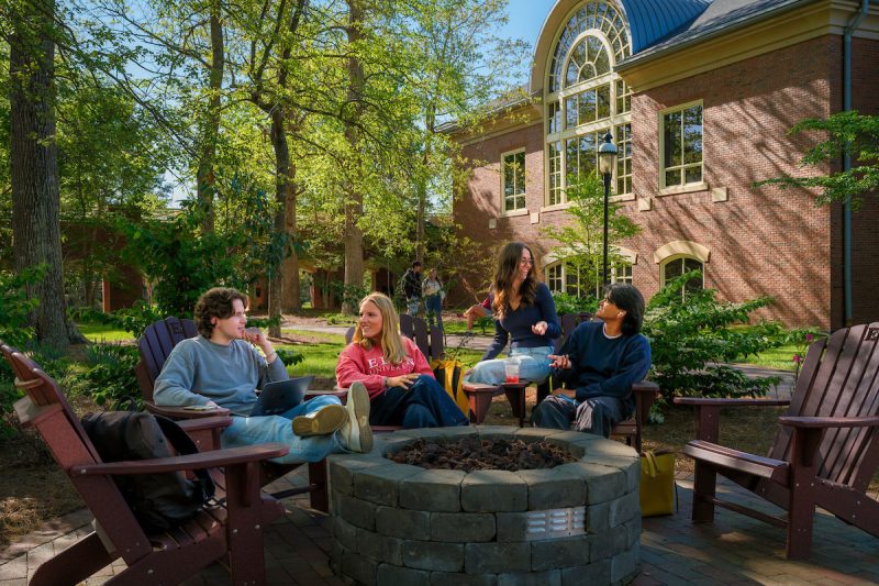 Four students gather around an outdoor fire pit, talking and studying on a sunny day outside a brick academic building.