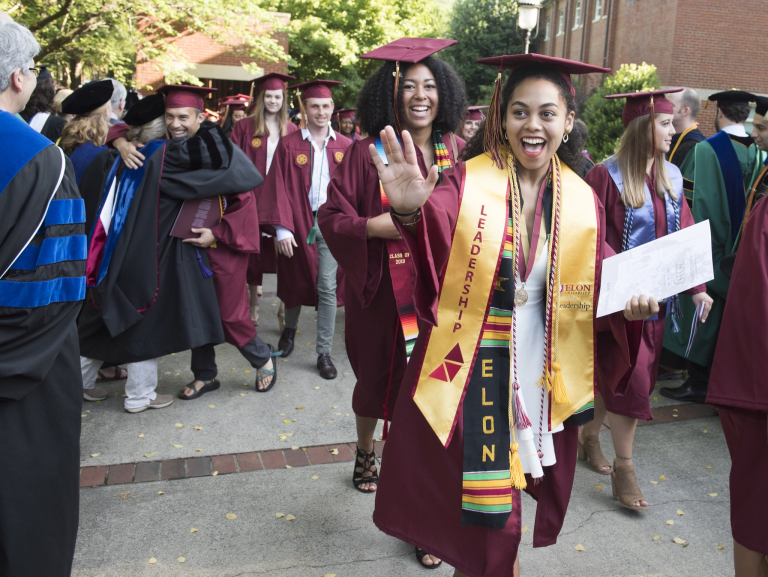 Elon graduates in maroon caps and gowns celebrate after the ceremony, smiling and waving as they walk to the venue.