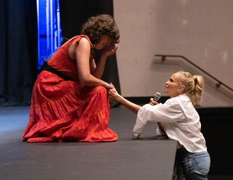 A student in a red dress kneels onstage while a professional actress, holding a microphone, speaks to her during a theater rehearsal or workshop.