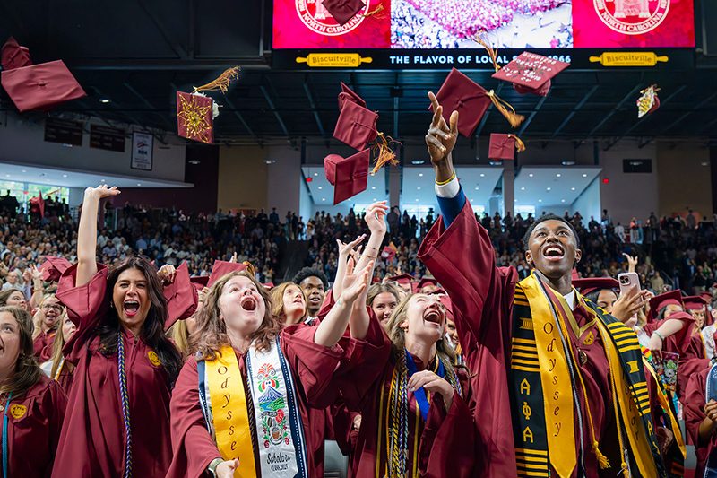 Graduates in maroon caps and gowns toss their hats in the air, celebrating at Elon University’s Commencement ceremony.