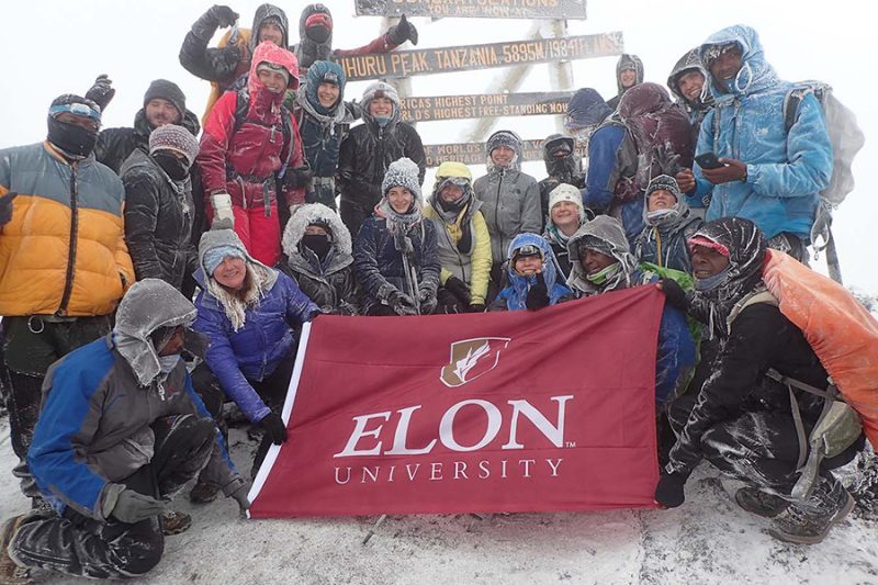 A group of students in winter coats stand at the snowy top of Mount Kilimanjaro holding a maroon Elon University banner.