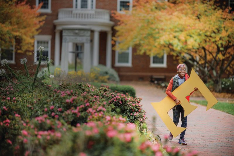 A student walks down a brick pathway on a college campus carrying a cardboard cutout in the shape of a giant capital letter E.
