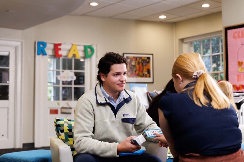 A teacher and student work together using flashcards in a reading classroom. The teacher listens attentively as the student reads a word card, with colorful “READ” letters and books visible in the background.