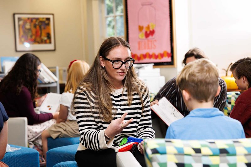 A teacher works one-on-one with a young student in a reading classroom, using colorful learning tools. Other students and teachers read and interact in the background, creating a warm, engaging learning environment.
