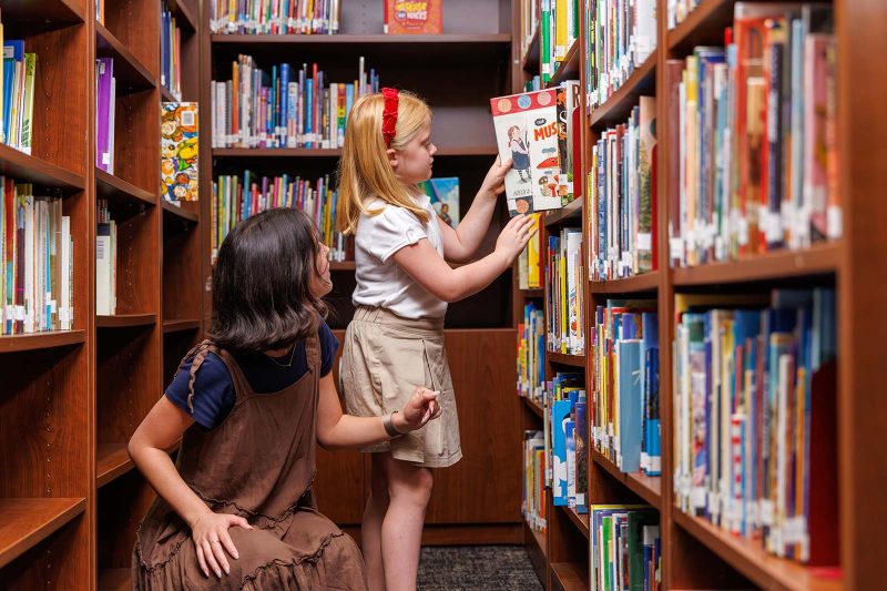 A young student selects a book from a library shelf while a teacher kneels nearby, offering guidance. Rows of colorful books fill the shelves, creating a bright and welcoming learning environment.