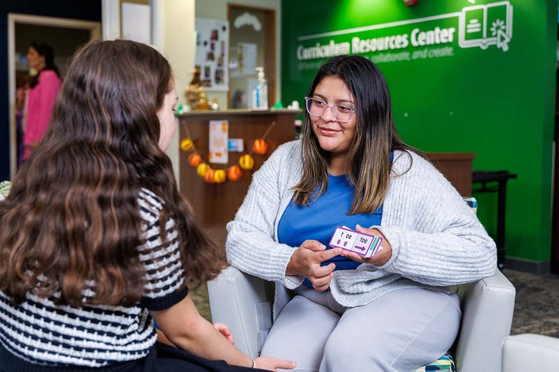 An educator works with a student in a reading classroom, holding flashcards to teach phonics. The two are seated in front of a green wall labeled “Curriculum Resources Center,” creating a bright and welcoming learning space.