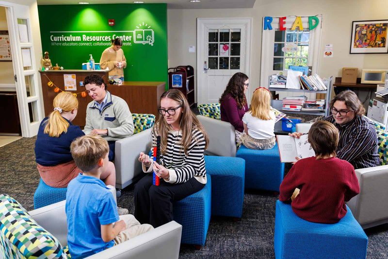 Teachers and students sit together at a round table, smiling as they work on a reading activity. The classroom is bright and colorful, filled with books, learning materials, and artwork that create a warm, supportive environment.