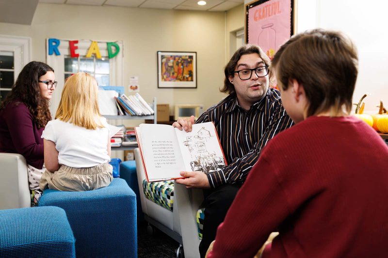 A teacher reads aloud from a picture book while engaging a student in discussion. Other students and teachers read together in the background, surrounded by books and colorful classroom decorations that spell the word READ.