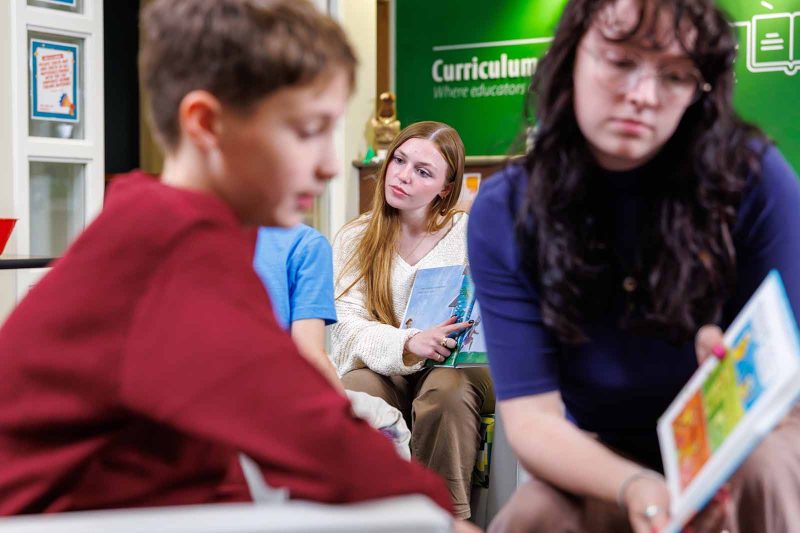 A teacher listens attentively as students participate in a reading activity. Another teacher in the foreground holds an open picture book, while children sit nearby in a bright, engaging classroom.