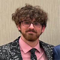 Jacob is wearing a suit jacket with a pink shirt and black tie. He has glasses and curly hair.