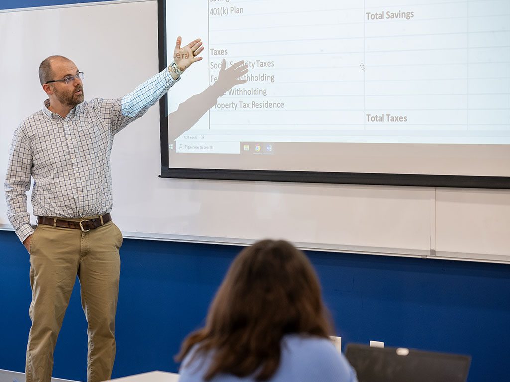 Photo of Chris Harris teaching a finance class while explaining a budgeting worksheet on a projected screen