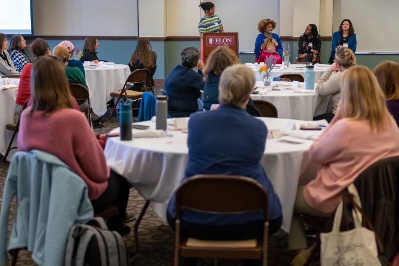 Attendees seated at round tables listen to a speaker at the Elon University podium during an International Women's Day event.