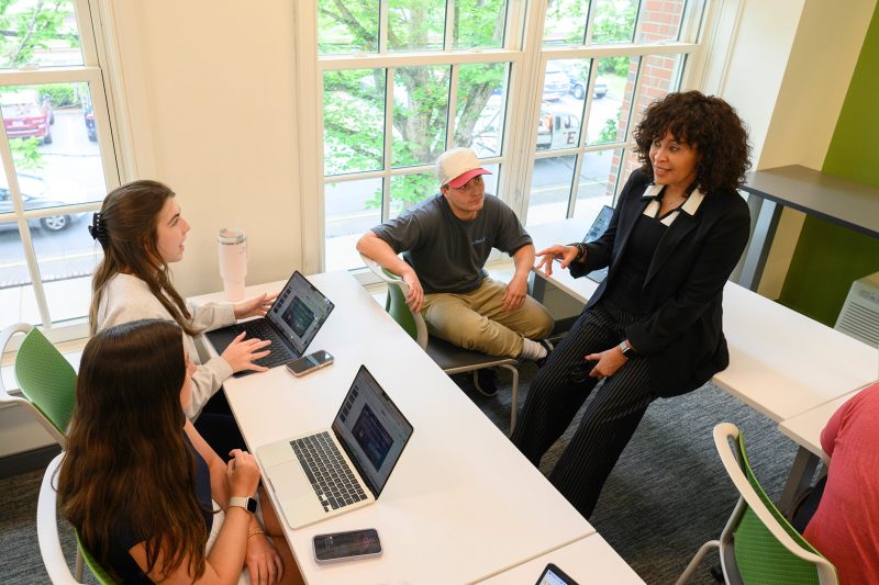 Faculty member leading a small group discussion with students around a table with laptops in a bright campus classroom.