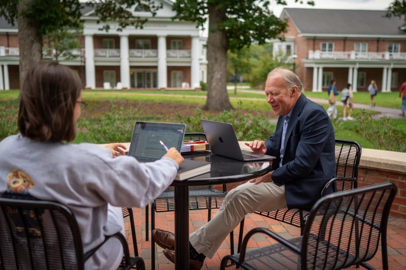 Photo of Brian Pennington, Director of the Center for the Study of Religion, Culture, and Society, meeting with a student outdoors at a table in the Lambert Academic Village at Elon University.