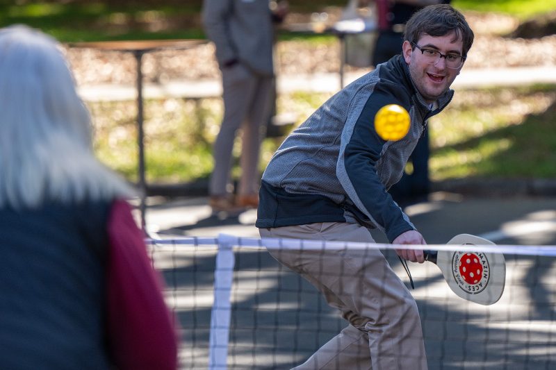 Elon University staff member smiling while playing pickleball during HealthEU Day, paddle in hand and ball in mid-air across the net.