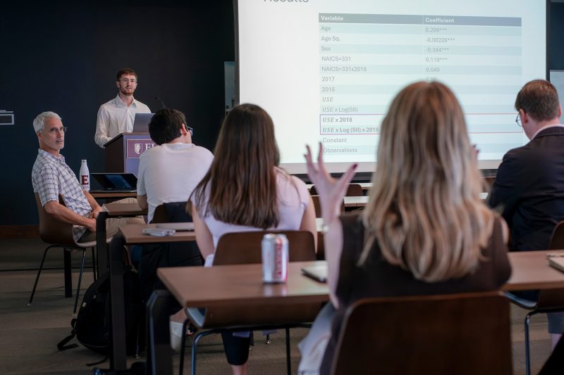 Elon University student presenting research results at an Elon podium during the Student Undergraduate Research Forum (SURF), with a regression coefficient table projected on screen and an audience member raising their hand.