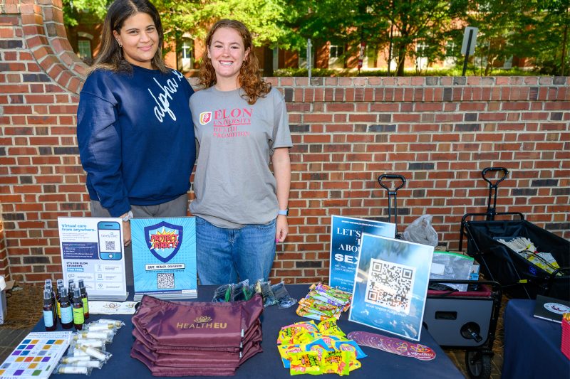 Two students staffing a HealthEU Day tabling event at Elon University, with health promotion materials, tote bags, and informational signs including TimelyCare and "Protect Direct" on display.