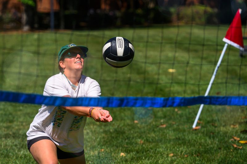Student bumping a volleyball at an outdoor grass court during a casual campus game on a sunny day.
