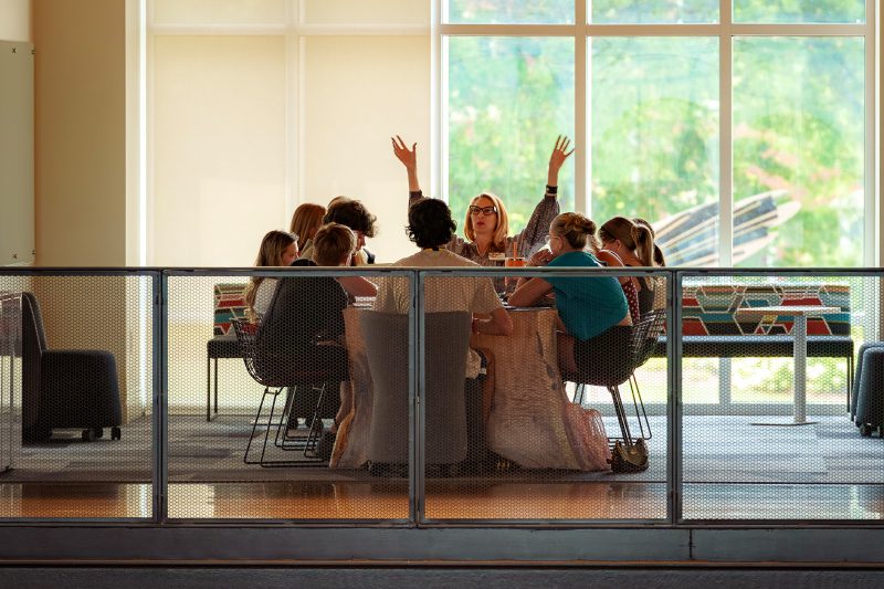 Elon faculty member leading a Poetic Light summer class around a table indoors, arms raised expressively, with students gathered closely in discussion.