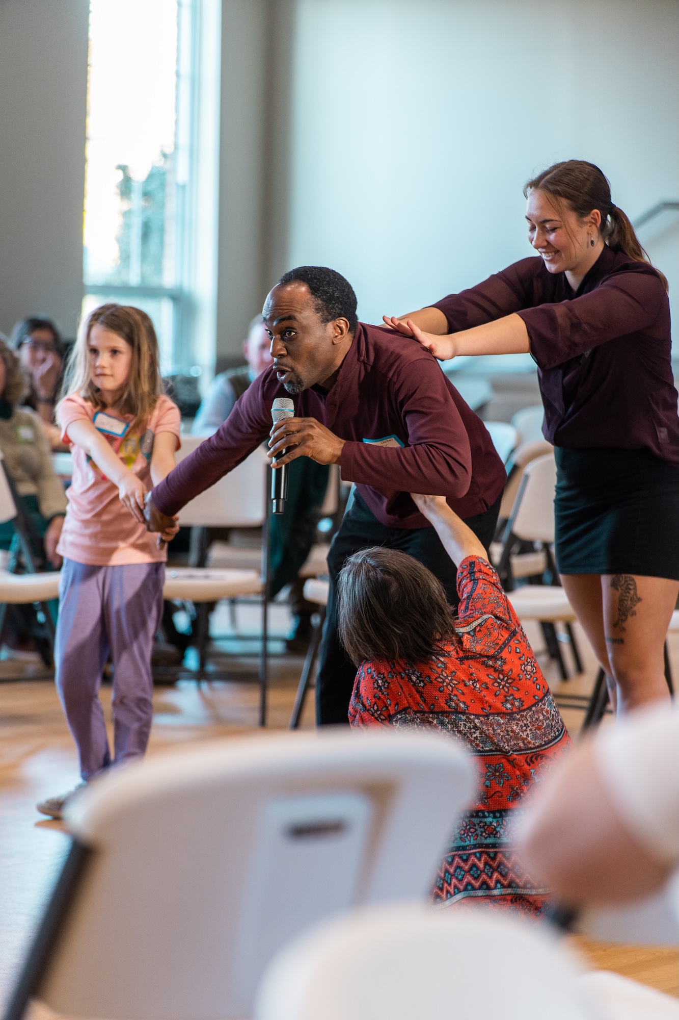 a workshop leader holding a microphone poses to demonstrate stuckness. A child pulls his hand and a student pushes his back to add to the sculpture