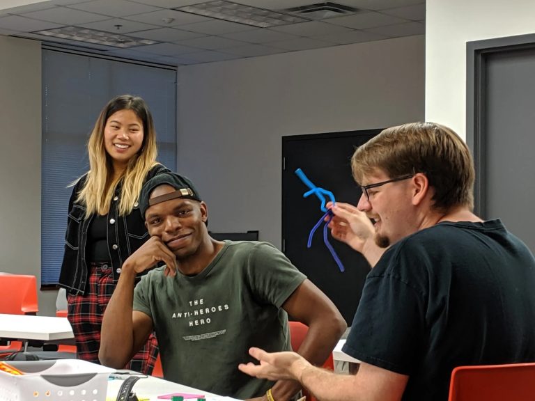 3 students are talking and laughing in the center for design thinking. The student in the middle is looking directly at the camera and smiling as the student on the right explains a pipe cleaner creation he is holding up.