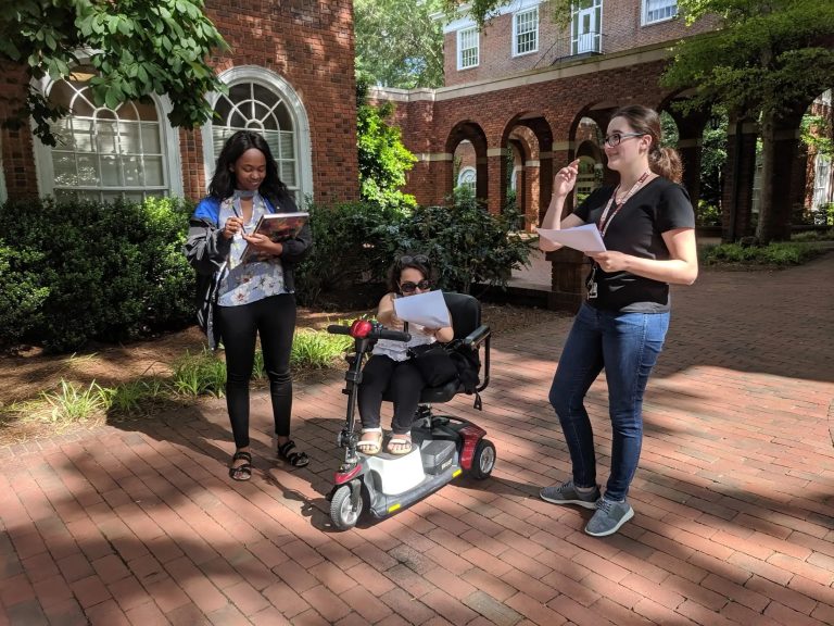 three students, one in a mobility scooter, are outside on Elon's campus. They are holding pens and paper to take notes about which places on campus are accessible to wheelchair and scooter users.