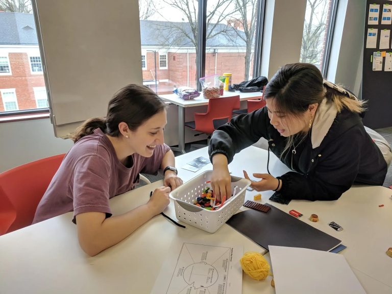 2 students look through a bucket of art supplies that sit in the middle of a work table. They are working on a creative project.