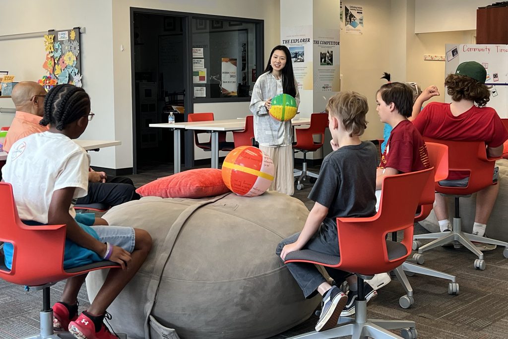 a masters of education student holds an inflatable beach ball with questions written on it. She is explaining a get-to-know-you game to young students as part of a teaching exercise