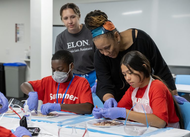 two elementary students sit a a table and examine cow intestines while an elon professor and an elon student look on and offer advice