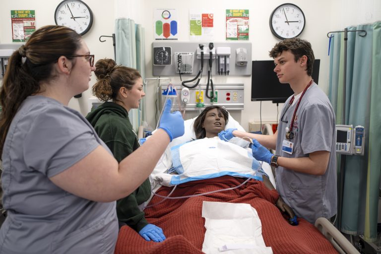 three nursing students stand around a patient mannequin which is lying in a hospital bed. The students are discussing how they might treat their mock patient in this instructional simulation