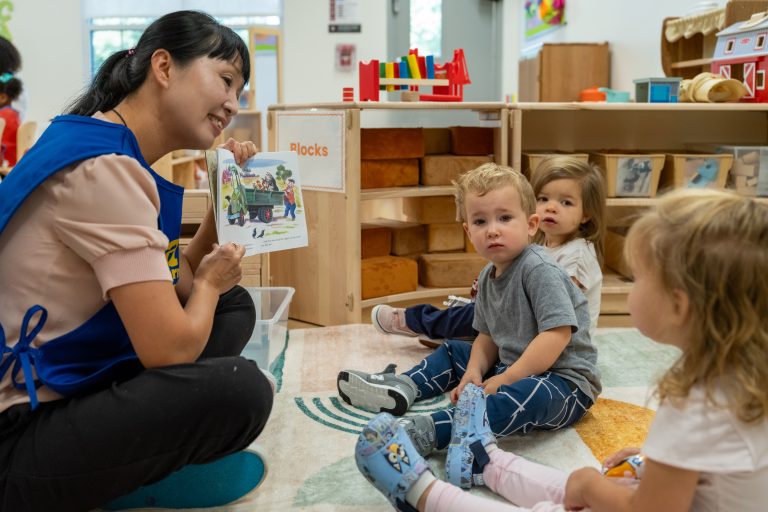 a teacher holds up a book she is reading so three toddlers sitting on the classroom floor can see the pictures